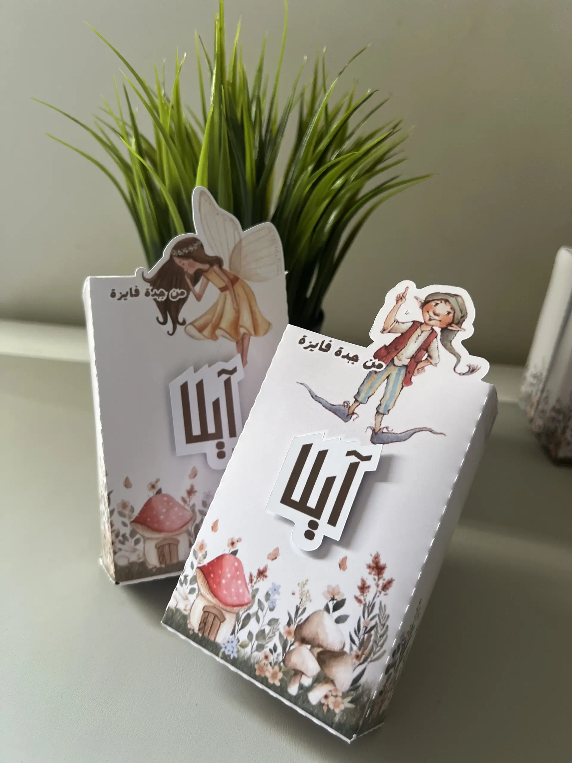 علب بيضاء برسوم فطر وزهور برية، يعلوها رسم جنية أو قزم مقصوص على حدوده، وعليها اسم الطفلة.